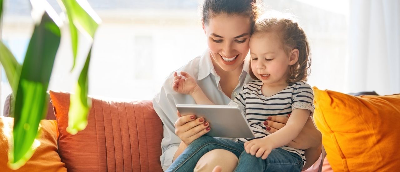 A mother and her young daughter smiling at a tablet on an orange sofa